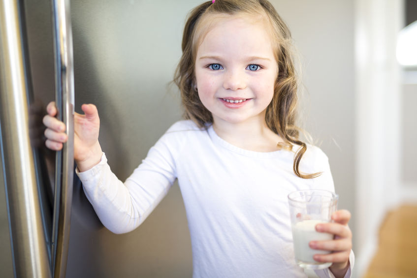 Young girl holding glass of milk at refrigerator.
