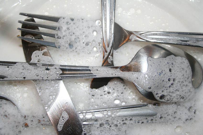 Silverware under running water in soapy sink.